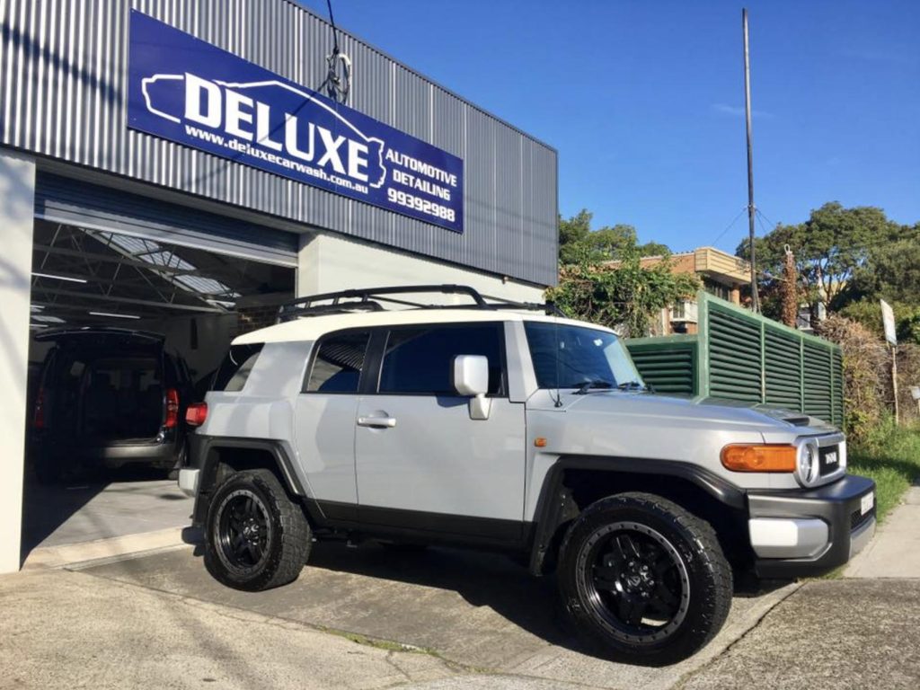 ### Key Elements  #### Main Subject - **Vehicle**: A silver Toyota FJ Cruiser parked outside a detailing shop.  #### Objects - **Shop Signage**: "DELUXE" prominently displayed, indicating the name of the automotive detailing business. - **Surroundings**: A garage-like structure with a clean, organized exterior and a green fence in the background.  #### Notable Features - **Vehicle Design**: Distinctive rugged appearance of the FJ Cruiser, with black wheels and a roof rack. - **Lighting**: Natural daylight enhances the vehicle's features and the shop's exterior.  #### Text/Symbols - **Shop Name**: "DELUXE" is the main text on the sign, indicating a focus on high-quality automotive services.  ### Overall Mood/Theme The image conveys a sense of professionalism and care in automotive detailing, with a focus on rugged, adventurous vehicles. The bright daylight and clean surroundings suggest a welcoming and well-maintained environment.