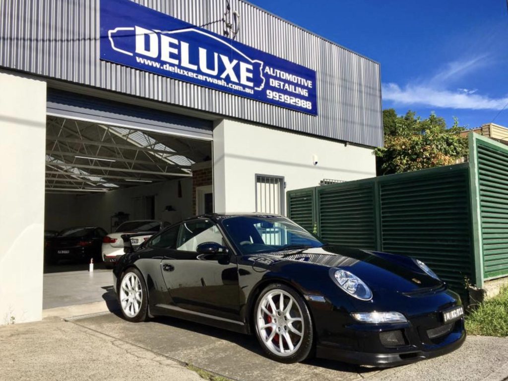 ### Key Elements  - **Main Subject**: Black sports car (Porsche) - **Notable Features**: Sleek design, white alloy wheels, polished appearance - **Background**: Automotive detailing shop with a clear sign - **Text/Symbols**: Sign reads "DELUXE AUTOMOTIVE DETAILING" with contact details - **Surroundings**: Other cars visible in the shop, greenery in the background  ### Overall Mood/Theme The image conveys a sense of luxury and professionalism, highlighting the meticulous care given to high-end vehicles. The bright lighting and clean environment suggest a focus on quality service and automotive aesthetics.