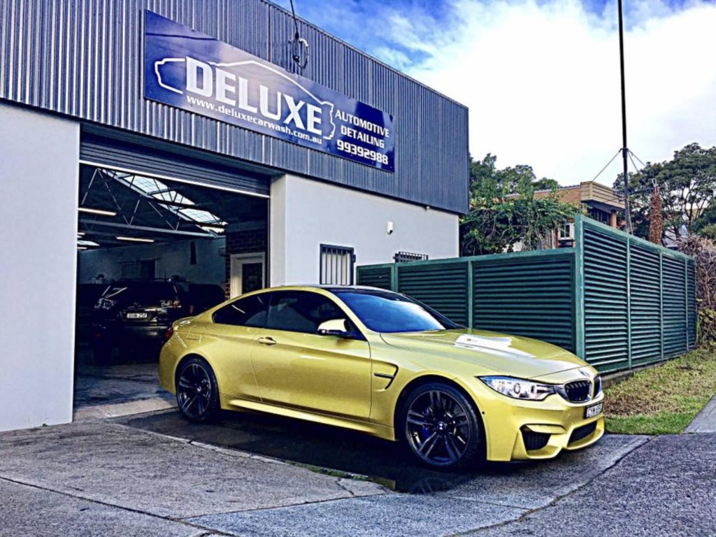 ### Key Elements  - **Main Subject:** A bright yellow car (BMW) parked outside a detailing garage. - **Garage Features:** The garage sign reads "DELUXE AUTOMOTIVE DETAILING" with visible contact information. - **Surroundings:** The garage has a modern design, with a grey exterior and a door partially open, indicating busy activity inside. - **Notable Objects:** Another car is partially visible inside the garage, suggesting additional services. - **Overall Mood:** The scene conveys a sense of professionalism and quality automotive care, highlighted by the striking color of the car and the neat presentation of the garage.