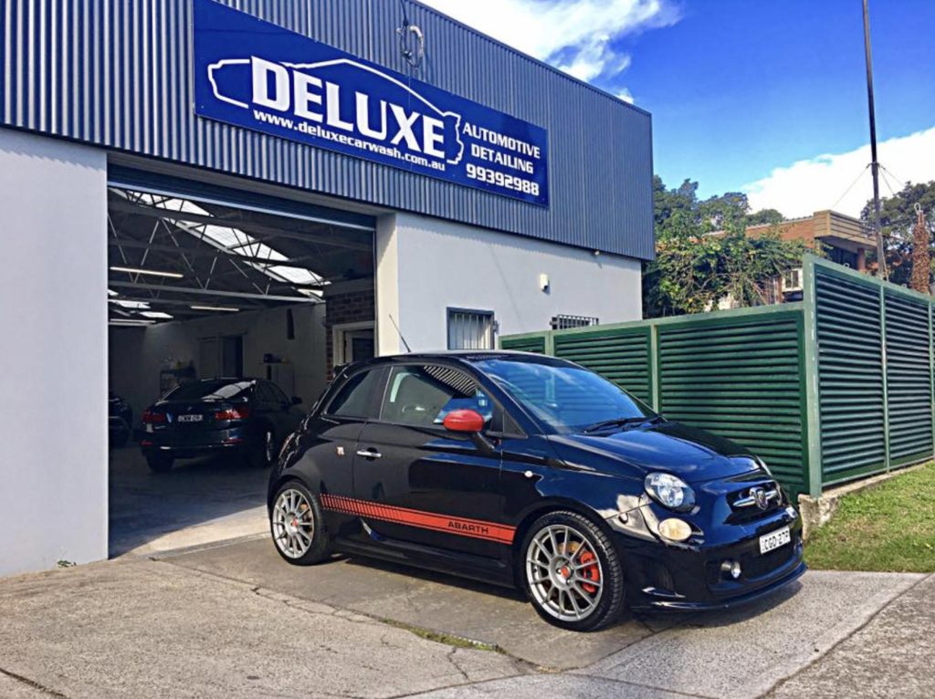 ### Key Elements  **Main Subject**   - A black Fiat 500 Abarth car.  **Surrounding Objects**   - A car wash facility named "DELUXE Automotive Detail" in the background. - Another vehicle visible in the background.  **Notable Features**   - The car features a distinctive red stripe and sporty wheels. - The car wash has a clean, modern appearance with a well-maintained exterior.  **Text/Symbols**   - The sign "DELUXE Automotive Detail" prominently displayed above the entrance.  ### Overall Mood/Theme   The image conveys a sense of professionalism and automotive care, highlighting a stylish, sporty vehicle in a well-maintained car wash environment. The setting suggests a focus on quality and attention to detail in car maintenance.